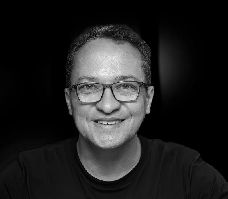 Retrato de Jackson Carvalho em preto e branco, de óculos e sorrindo, sobre fundo preto