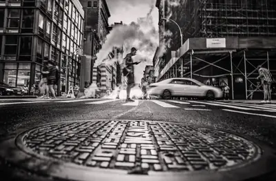 Rua de Nova York com uma tampa de bueiro em primeiro plano, vapor saindo do asfalto e prédios ao fundo
